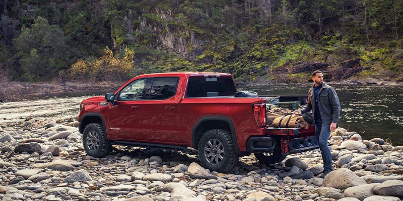 A red 2026 GMC Sierra 1500 truck is parked on the mountain creek shore of quite large rocks with the driver removing camping equipment from the opened truck bed.