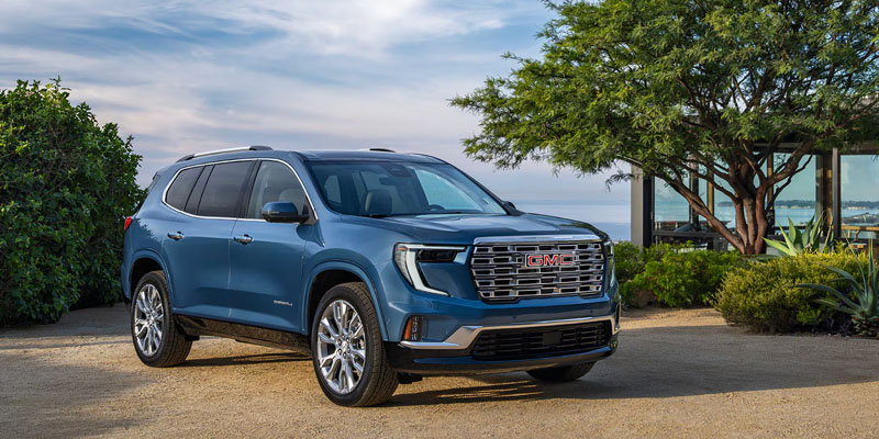 A deep blue sky-colored 2026 GMC Acadia sits in perfect harmony beside a large blue lake or seashore with green trees, bushes and a white cloud-laced sky behind.