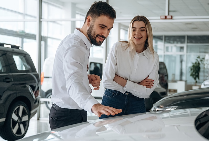 A smiling white-shirted couple pat the shining surface of a GMC vehicle on the sunlit showroom floor.